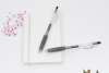兩支透明原子筆擺放在白色硬皮筆記本上的情境照。背景簡約，左上角裝飾有一張粉紅色櫻花圖案的卡片，營造出清新專業的文具氛圍。