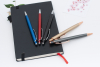 另一角度的情境照，藍、紅、黑三支筆靠在黑色筆記本邊緣，香檳金與鐵灰筆則交叉平放於前方，強調了鋁合金筆身細長的流線造型。