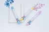 三支不同粉彩色系（淺藍、淡黃、粉紅）的六色原子筆，筆身印有淺色幾何圖形，整齊擺放在白色筆記本與藍色、粉紅花卉裝飾卡片旁。