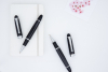 兩支黑色鋼珠筆擺放在白色硬皮筆記本上的情境照。一支筆蓋已取下放置在旁，露出銀色的錐形筆尖與具有防滑橫紋設計的黑色握把，背景點綴著粉紅色的櫻花裝飾卡片。