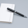 一支黑銀金屬原子筆斜放在白色硬皮筆記本封面上的情境照。背景為純淨的灰色調，突顯出產品作為高端辦公文具的專業質感。