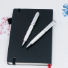 兩支白色金屬原子筆交叉擺放在黑色硬皮筆記本上的情境照。背景點綴著清新風格的櫻花與藍色圖騰卡片，營造出高雅且具專業感的辦公與書寫氛圍。