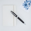 一支 Lensen 金屬原子筆斜放在白色硬皮記事本上的情境照。背景右上角配有一張帶有藍色花卉圖案的裝飾卡片，展現出簡潔且具質感的商務書寫氛圍。