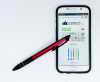 一支紅色三色筆擺放在智慧型手機旁，手機螢幕顯示購物網站，強調其電容觸控頭可操作螢幕的功能。